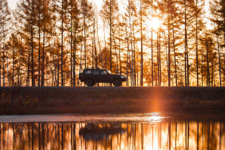 Black SUV on foreat road near a lake at sunsetの写真素材