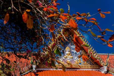 The roof of a Buddhist temple with the image of the Buddha and blooming trees against the blue sky. Buddhist temples in Thailand. Religion of Southeast Asia. Tourist destination of Thailand.の写真素材