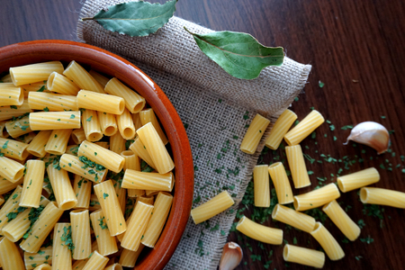 bowl of round clay filled with macaroni and parsley, on top of a linen napkin, garlic and macaroni spilled on the wooden table.の写真素材