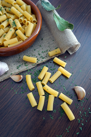 bowl of round clay filled with macaroni and parsley, on top of a linen napkin, garlic and macaroni spilled on the wooden table.の写真素材