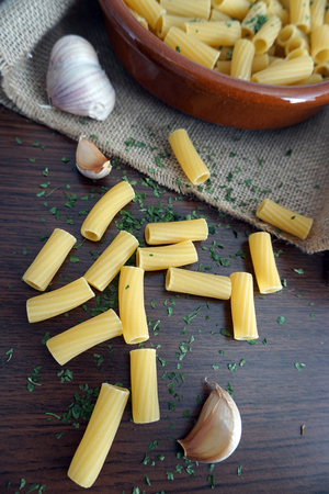 bowl of round clay filled with macaroni and parsley, on top of a linen napkin, garlic and macaroni spilled on the wooden table.の写真素材