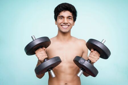 Young handsome asian boy holding dumbbells on both the hands looking into the camera while smiling. Standing in front of a blue wall.の写真素材