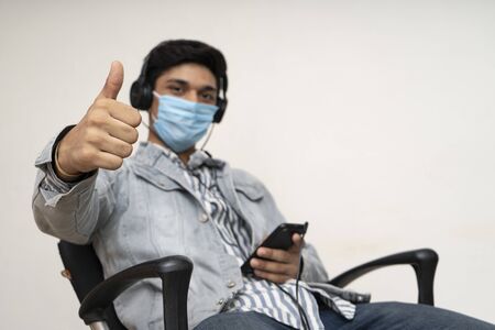 Young handsome asian indian boy sitting on a chair, wearing a protective mask, showing thumbs up in the camera.の写真素材