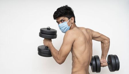 Fitness man with mask for epidemic protection working exercise for arm,biceps, and shoulders with dumbbells.Training in epidemic timeの写真素材