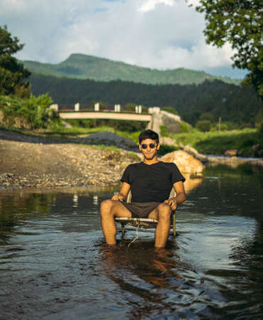 A young handsome asian boy sitting in a chair in the middle of a river with black sunglasses, golden hour.の写真素材