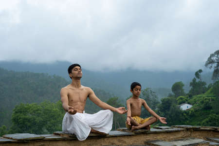 A young handsome boy doing yoga with a kid on the roof of a house situated in the middle of mountain range. Yoga and fitness.の写真素材