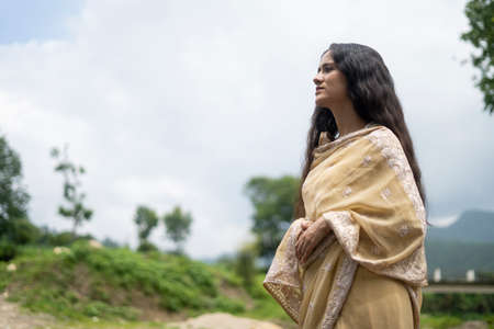 Beautiful Indian young female model wearing traditional saree standing in front of a river landscape with overcast clouds.の写真素材