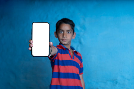 young indian kid holding a phone with white screen, standing against a blue background.