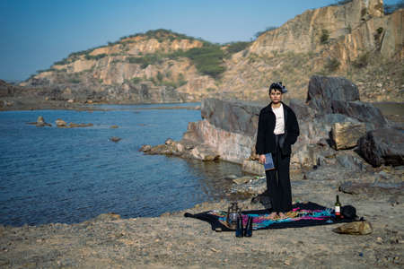 Young beautiful indian girl reading a book sitting on a blanket in the mountains with a lake nearby....