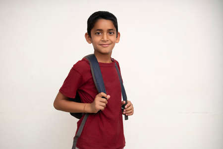 Young indian kid standing on a white wall with a school bag on his back. Getting ready for school. School and education concept.の写真素材