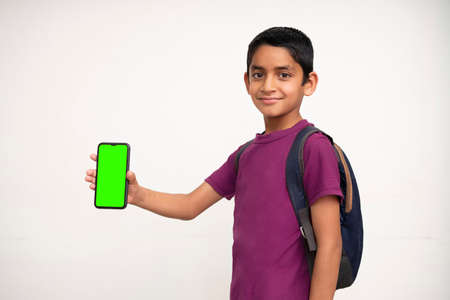 Young Indian kid holding a phone in his hand with a green screen, school bag in his back and wearing purple t-shirt standing on white isolated background.の写真素材