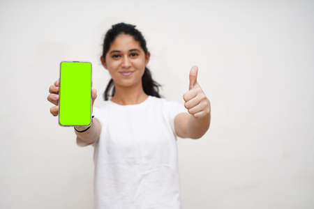 Young indian beautiful girl in white dress and white background holding a mobile phone with green...