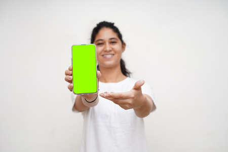 Young indian beautiful girl in white dress and white background holding a mobile phone with green screen used for mockups of mobile. Mobile and technology concept.の写真素材