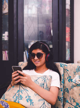 Young beautiful girl sitting on a sofa and using her mobile phone while listening to songs on her...