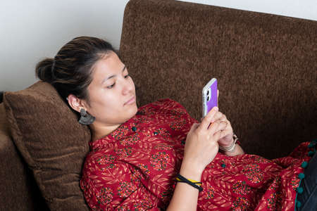 Young indian girl using her mobile phone while lying down on the couch.Lifestyle and technology concept.の写真素材