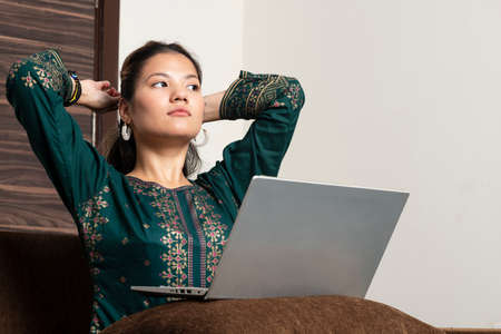 Young indian girl working on her laptop while lying down on the couch.smiling hindu female professional employee typing email on laptop at workplaceの写真素材