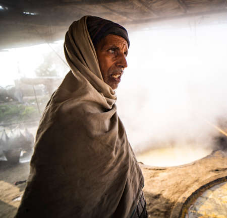 Garh Mukteshwar, Uttar Pradesh- January 13 2022 - Portrait of an old aged man in a factory. Old aged man in the village with turban and with smoke in the background.のeditorial素材