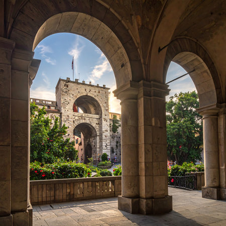 View of a historical city gate, framed by arches, in a bustling European city square during a pleasant summer day. Stone archways provide a perspective of a beautifully preserved medieval gate and surrounding courtyard, with trees and gardens. The light suggests a warm, sunny day.の素材