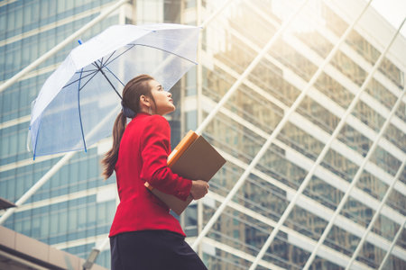 Young beautiful Asian business woman holding umbrella and documents walking to office for working under sunlight and hot weather.の写真素材