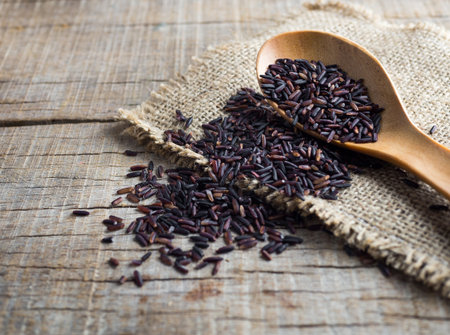 Close up rice berry(brown rice, Thai black jasmine rice or raw purple/red rice) in a wooden bowl, spoon and sack on wooden background.の写真素材