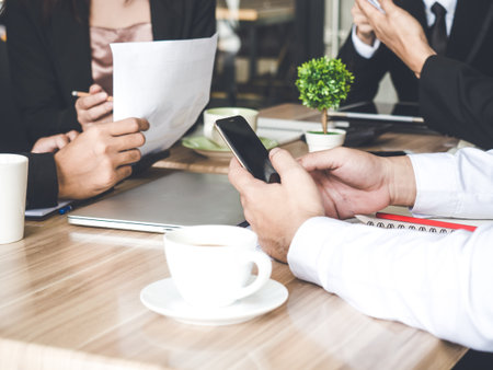 Meeting table/desk, businessman using smart phone and checking business plan, progress report for business work, financial report, status, tablet, coffee, on table, discussing with colleague at officeの写真素材