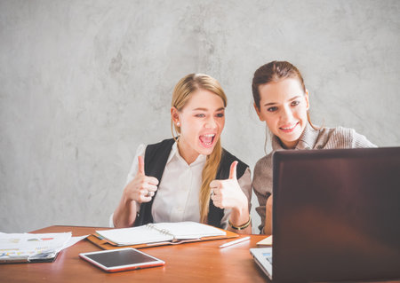 Two of young caucasian business woman working with happy feeling and smile in the office, sitting on desk with computer laptop reading good news and feeling surprise.の写真素材