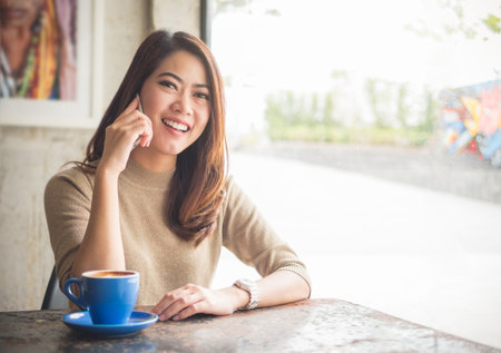 Young beautiful asian woman using smart phone talking with friends for business in coffee shop over the blurred background, happy and smile feelingの写真素材