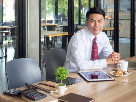 Portrait young asian businessman working with tablet, computer, documents and coffee on table at cafe office, casual business/businessman in the morning happy feeling conceptの写真素材