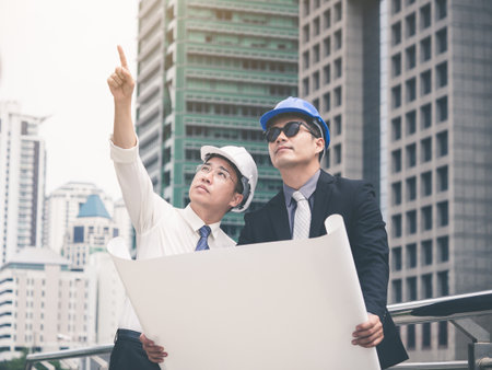 Young asian engineer/architect holding drawing/blueprint discuss with businessman for a project construction city building, happy feeling, positive, teamwork and togetherness conceptの写真素材