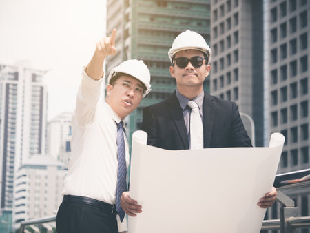 Young asian engineer/architect holding drawing/blueprint discuss with businessman for a project construction city building, happy feeling, positive, teamwork and togetherness conceptの写真素材