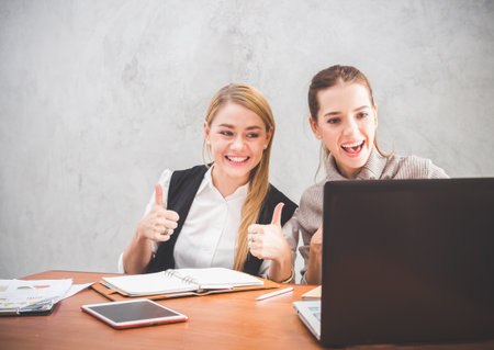 Two of young caucasian business woman working with happy feeling and smile in the office, sitting on desk with computer laptop reading good news and feeling surprise.の写真素材