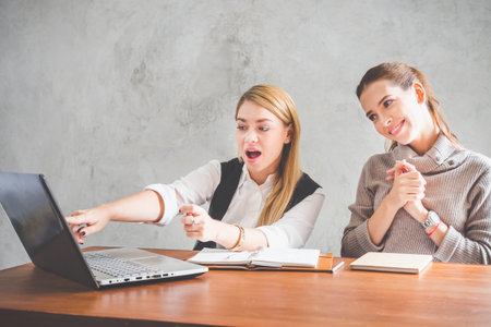 Two of young caucasian business woman working with happy feeling and smile in the office, sitting on desk with computer laptop reading good news and feeling surprise.の写真素材