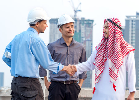 Young asian engineer/architect shaking hands with arab businessman, talking discussing about a project construction building, teamwork, togetherness, positive feeling conceptの写真素材