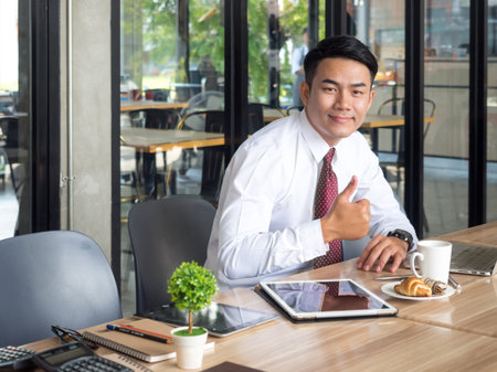 Portrait young asian businessman showing thumbs up while working with tablet, computer, documents and coffee on table at cafe office, casual business/businessman in the morning happy feeling conceptの写真素材