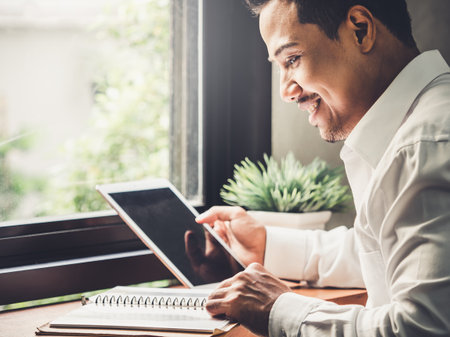 Young asian business man working on tablet with happy smile and good mood, sitting over window with sunlight reading news and financial report, vintage toneの写真素材