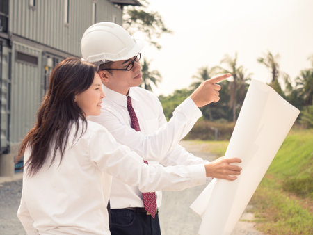 Young engineer/architect pointing to construction building plan to customer, hand holding drawing blueprint plan for home/factoryの写真素材
