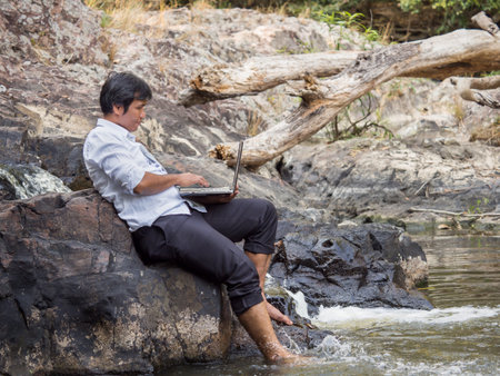 Middle age Asian businessman wears casual suit working in nature forest with labtop computer, working with relaxing and chilling lifestyle, happy for working business in forest conceptの写真素材