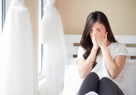 Young beautiful asian woman headache on white bed, sad, stressed, crying, disappointed feeling in the morningの写真素材