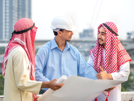 Young asian engineer/architect holding drawing/blueprint discuss with arab businessman for a project construction building, happy feeling, positive, teamwork and togetherness conceptの写真素材