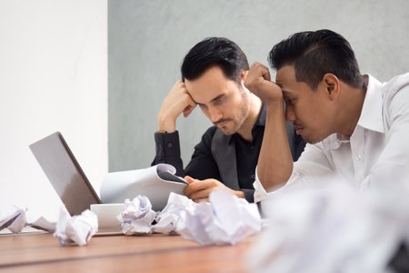 Two of young caucasian and asian businessman upset tired from work sitting at desk in office, unemployment, fired from job, disappointed, loss and feeling down conceptの写真素材