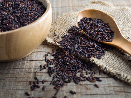 Close up rice berry(brown rice, Thai black jasmine rice or raw purple/red rice) in a wooden bowl, spoon and sack on wooden background.の写真素材