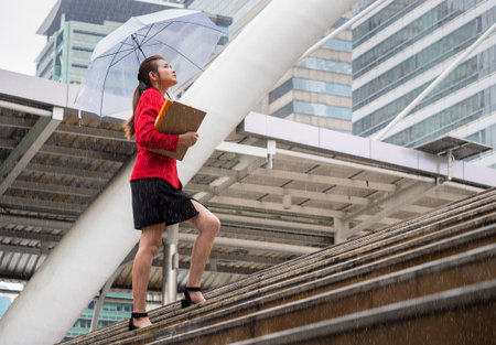 Young asian businesswoman holding business documents and umbrella walking under raining in the cityの写真素材