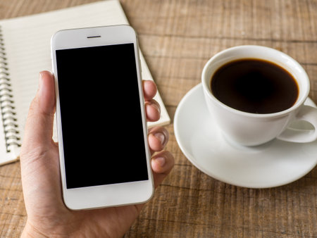Close up business man's hand holds smart phone with black isolated screen over background of notebook, coffee on wooden backgroundの写真素材
