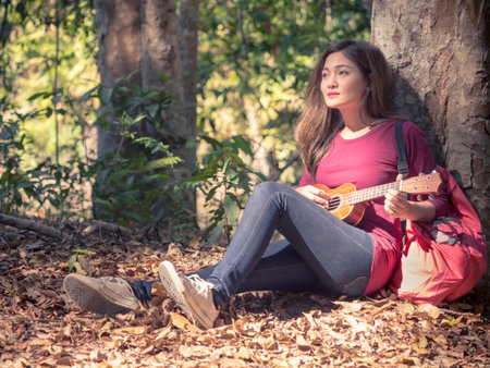 Young beautiful asian woman sitting at tree playing ukulele/guitar and sing a song chilling at forest nature during travle outdoor camping/trekking with happy feeling/relaxing conceptの写真素材