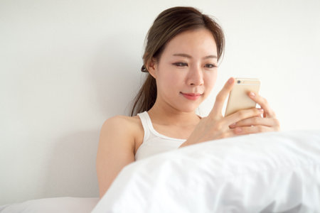 Young asian woman wake up happy, using smart phone reading good news on the bed with fresh feeling in the morningの写真素材