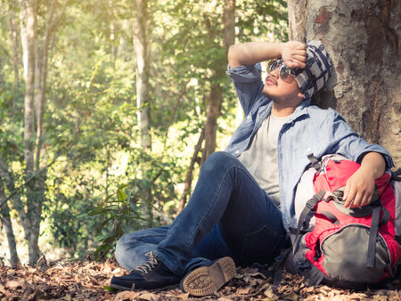 Young asian man wears jeans(hipster style) with backpack sitting at tree (take a rest) in nature forest during trekking/hiking/traveling on vacation/holiday, freedom lifestyle outdoor forest conceptの写真素材