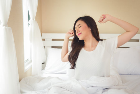 Asian woman wake up in the morning, sitting on white bed and stretching, feeling happy and freshの写真素材