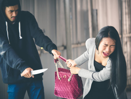 Culprit on hood holding knife is stealing bag from asian woman while walking alone at street, danger, burglar, criminal and robbery for woman concept.の写真素材