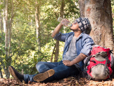 Young asian man wears jeans(hipster style) with backpack sitting at tree (take a rest) in nature forest during trekking/hiking/traveling on vacation/holiday, freedom lifestyle outdoor forest conceptの写真素材