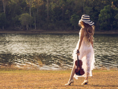 Young woman musician/violinist on white dress holding violin over river/park/forest, freedom lifestyle music vintage tone concept.の写真素材